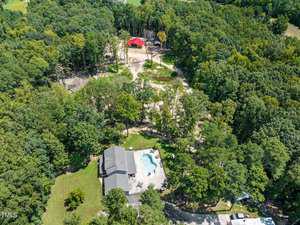 Aerial view of a wooded area with a large house and swimming pool surrounded by trees. There's a dirt path leading to a red-roofed building in the background. The scene is lush and green, indicating a rural setting.
