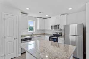A modern kitchen with white cabinets, granite countertops, a central island, and stainless steel appliances including a refrigerator, oven, and dishwasher. A window above the sink lets in natural light.