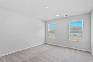 An empty room with light gray walls and carpeted flooring. Two windows on one wall allow natural light, showing an outdoor grassy area and blue sky. The ceiling has recessed lighting.