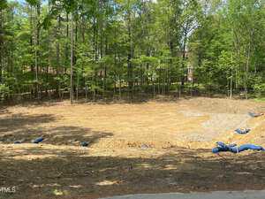 A cleared patch of land surrounded by trees, featuring a large, leveled dirt area with blue erosion control barriers on the ground. The site is bordered by a dense forested area under bright sunlight.
