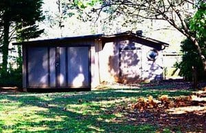 A small, simple shed with a flat roof stands in a grassy yard surrounded by trees. Dappled sunlight filters through the leaves, creating shadows on the shed and lawn. A pile of fallen leaves is visible in the foreground.