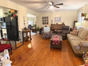 A cozy living room with wooden floors, featuring a sofa, recliner, and a coffee table with a red runner. A ceiling fan is above, and a kitchen is visible in the background. Various decorations, plants, and a piano add charm to the space.