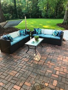 A brick patio with two wicker sofas featuring blue cushions and patterned throw pillows. A glass-topped coffee table is placed between the sofas. Lush greenery and trees are visible in the background.