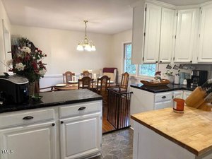 A cozy kitchen and dining area with white cabinets and black countertops. The dining room features a wooden table with chairs under a hanging light. A safety gate is visible near the kitchen entrance, and a window lets in natural light.