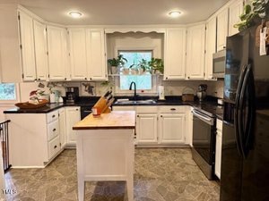 A kitchen with white cabinets, black countertops, and a central wooden island. A window above the sink has potted plants on the sill. There are modern appliances, including a stainless steel oven and a black refrigerator.