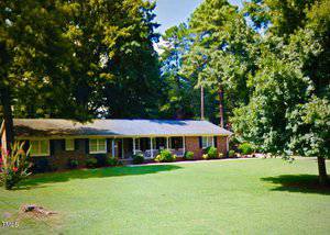 A single-story brick house with a covered porch sits surrounded by lush green grass and tall trees. Bushes and flowerbeds line the front, and the sky is clear and sunny.
