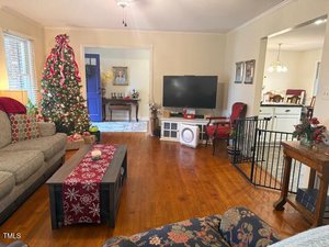 A cozy living room decorated for Christmas, featuring a Christmas tree with red and silver ornaments, a TV, a couch, and a coffee table with a red patterned runner. A baby gate separates the living area from the adjacent kitchen.