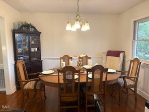 A dining room with a wooden table set for six, featuring cream-colored plates and candles. A cushioned chair sits by a window with a view of trees. A dark wooden china cabinet is in the corner, under a chandelier with warm lighting.