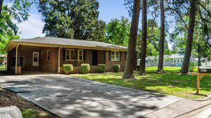 A charming single-story brick house at 227 W Dale Drive in Oxford features a covered front porch and large windows, nestled among trees. A concrete driveway meanders to the home, with a mailbox labeled "1287" gracing the lawn's edge under a clear, sunny sky.