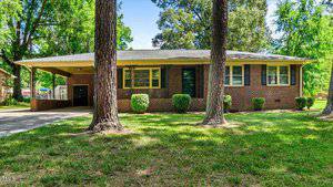 A single-story brick house with a carport attached, surrounded by trees and a well-kept lawn. The windows have yellow shutters, and there are small bushes lining the front of the house.