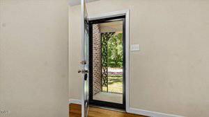 Open front door revealing a view of a brick porch and green, wooded yard. The beige interior walls and wooden floor of the house are visible. Natural light illuminates the entryway.