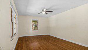 A vacant room with light-colored walls and hardwood flooring. Two windows on adjacent walls allow natural light in. A ceiling fan with a light fixture is centered on the ceiling.