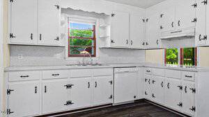 A bright kitchen with white cabinets, black handles, and gray speckled countertops. There's a sink under a window showing greenery outside. A dishwasher is installed next to the lower cabinets, and a range hood is mounted above the stovetop area.