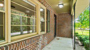 A small porch with brick walls, a tiled roof, and a concrete floor. Large windows with cream-colored frames are on the left. Surrounded by greenery, the porch features a black exterior light.