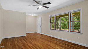 A vacant room with beige walls, a ceiling fan, and wooden floors. A door is on the left wall, and three windows on the right provide a view of greenery outside.