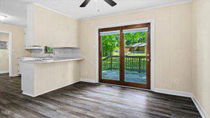 A bright room with light-colored walls and wood flooring. Double glass doors lead to a wooden deck overlooking a grassy backyard. A ceiling fan is above, and the kitchen area with a counter is visible to the left.