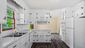 A bright kitchen featuring white cabinets with black handles, a white oven, and a double sink under a window. The room has dark wood flooring and light-colored walls, creating a fresh and clean atmosphere.