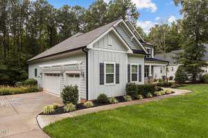 A modern suburban house with light gray siding, a dark roof, and white trim. It features a two-car garage, well-maintained bushes, a grassy lawn, and a backdrop of tall trees under a partly cloudy sky.