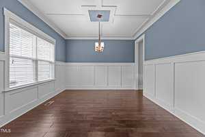 A spacious dining room with blue and white wainscoted walls, a hardwood floor, and a simple ceiling light fixture. A large window on the left lets in natural light.