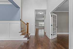 Interior of a modern home featuring a wooden staircase on the left, hardwood floors throughout, white walls, and a room with French doors on the right. A hallway leads to another room with large windows at the far end.