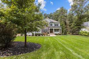 A two-story house with a stone and siding exterior, surrounded by a lush green lawn and various trees. The sky is clear with a few clouds, and the setting appears peaceful and well-maintained.