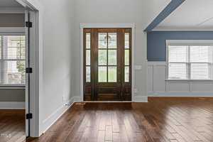 A bright foyer with a wooden front door featuring glass panels, opening into a room with hardwood floors. The walls are light gray with white trim, and a large window allows natural light into the space.