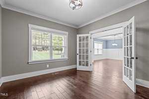 A spacious, empty room with light gray walls, hardwood floors, and a large window with blinds. French doors open to an adjacent room with similar design, both featuring modern overhead lights. Natural light fills the space, highlighting the clean interior.