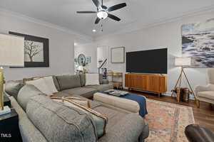 A cozy living room with a gray sectional sofa, a wooden mid-century modern TV stand with a flat-screen TV, two lamps, and landscape artwork on light walls. The room features a ceiling fan and a patterned rug on wooden flooring.