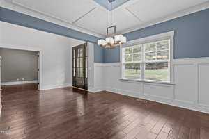 A modern dining room featuring light blue walls with white wainscoting. It has a dark wood floor and a white coffered ceiling with a rectangular chandelier. Large windows allow natural light to fill the space.