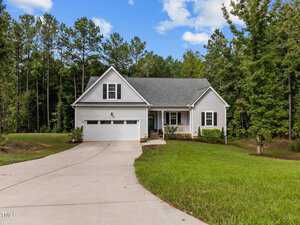 Nestled at 2150 Emerald Lane in Franklinton, this charming single-story gray house features a white garage and is surrounded by mature trees. A concrete driveway leads to the front porch adorned with white railings, while a well-manicured lawn complements the partly cloudy sky above.