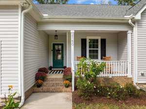 A cozy white house with a front porch. Two wooden chairs are set to the right near a black door. Potted plants and colorful flowers line the entrance, and a house number "2150" is displayed next to the door. Lush shrubs grow in the garden bed.