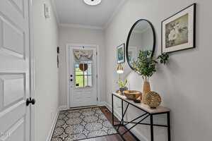 A hallway with a decorative rug, a console table with plants and a gold bowl, a round mirror on the wall, and framed artwork. A glass door leads to an outside view, allowing natural light in. The walls are painted light gray.