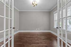 An empty room with light gray walls and dark hardwood floors, featuring a modern hanging light fixture. The room has French doors and two windows on the right, allowing natural light to enter.