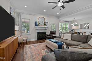 A cozy living room with a gray sectional sofa and patterned rug facing a large TV on a wooden stand. A fireplace is centered on the back wall, flanked by windows. Ceiling fan above, and a glimpse of the dining area in the background.