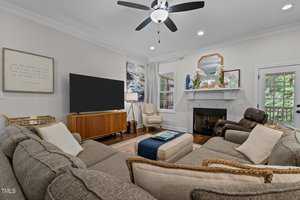 A cozy living room with a gray sectional sofa, a wooden TV stand with a large flat-screen TV, framed art on the walls, a fireplace with decor, an armchair, and a ceiling fan. A door leads outside, with greenery visible through the windows.