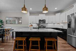 Modern kitchen with a large island featuring a marble countertop and three wooden stools. White cabinetry contrasts with black base cabinets. Stainless steel appliances, including a fridge and stove, and two gold pendant lights hang above.
