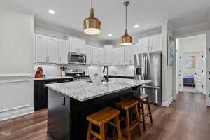 A modern kitchen featuring a black island with a white marble countertop and three wooden stools. It has white upper cabinets, stainless steel appliances, and two gold pendant lights. A hallway is visible in the background.