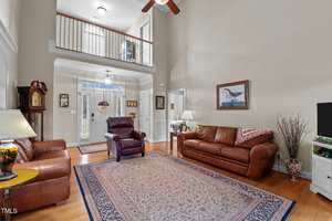A living room with high ceilings and hardwood floors features leather couches, a plush chair, a decorative rug, a grandfather clock, and a TV. A framed picture hangs on the wall, and the entryway with a window above the door is visible.