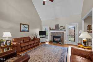 A cozy living room with high ceilings, leather sofas, and a patterned rug. A fireplace with decorative tiles is flanked by a door and a window. The room is warmly lit by lamps, and a ceiling fan hangs above. A painting and photos adorn the walls and tables.