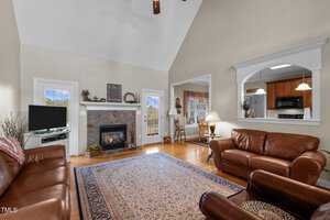 A cozy living room with high ceilings features a fireplace, a TV, and brown leather sofas. A patterned rug covers the wooden floor. A dining area and a kitchen with wooden cabinets are visible in the background. Plants and decor add warmth to the space.