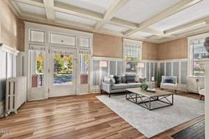 A spacious living room with large windows letting in natural light. It features a coffered ceiling, wood-paneled walls, and hardwood floors. The room is furnished with a gray sofa, a black coffee table, and a light gray area rug.