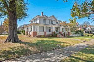 A large, two-story white house with a wraparound porch and brick foundation, surrounded by a grassy lawn and trees. The sky is clear and blue. The sidewalk runs alongside the home.
