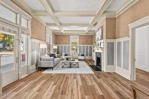 A spacious living room with large windows, a coffered ceiling, and hardwood floors. It features a gray sofa, two armchairs, a glass coffee table, and a fireplace. Natural light fills the room, highlighting the neutral-toned walls and decor.