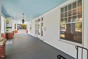 A spacious wooden porch with white siding, blue ceiling, and large windows. A black lantern hangs from the ceiling, and a mat lies near the entrance. Brick accents line the porch's edge, and trees are visible in the background.