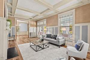 Elegant living room with hardwood floors, coffered ceiling, and large windows. Features a gray sofa, armchair, and a coffee table on a light rug. A fireplace stands to the left, and an open doorway leads to a dining area with a chandelier.