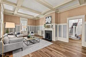 A spacious living room with light gray walls and wooden floors. It features a gray sofa, a glass coffee table, and a fireplace. Large windows with white trim let in natural light, and a staircase is visible in the background.