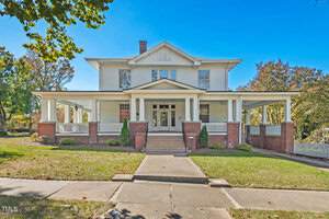A large, two-story white house at 316 Main Street boasts a spacious wraparound porch with brick columns and steps leading to a double-door entrance. Nestled in Oxford, the yard is neatly landscaped, surrounded by trees under a clear blue sky.