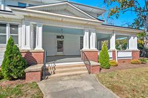 A large, white, two-story house with a gabled roof and a spacious front porch. The porch features red brick columns with white railings, flanked by small shrubs on either side of the steps leading to the entrance.