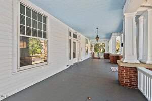 A spacious porch with light blue ceiling, white siding, and dark gray flooring. It features large windows, wooden doors with glass panels, a hanging light fixture, and red brick accents at the far end with a railing.