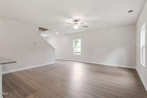 Empty living room with light wood flooring, white walls, and a ceiling fan. A staircase is visible on one side, and there's a large window letting in natural light.